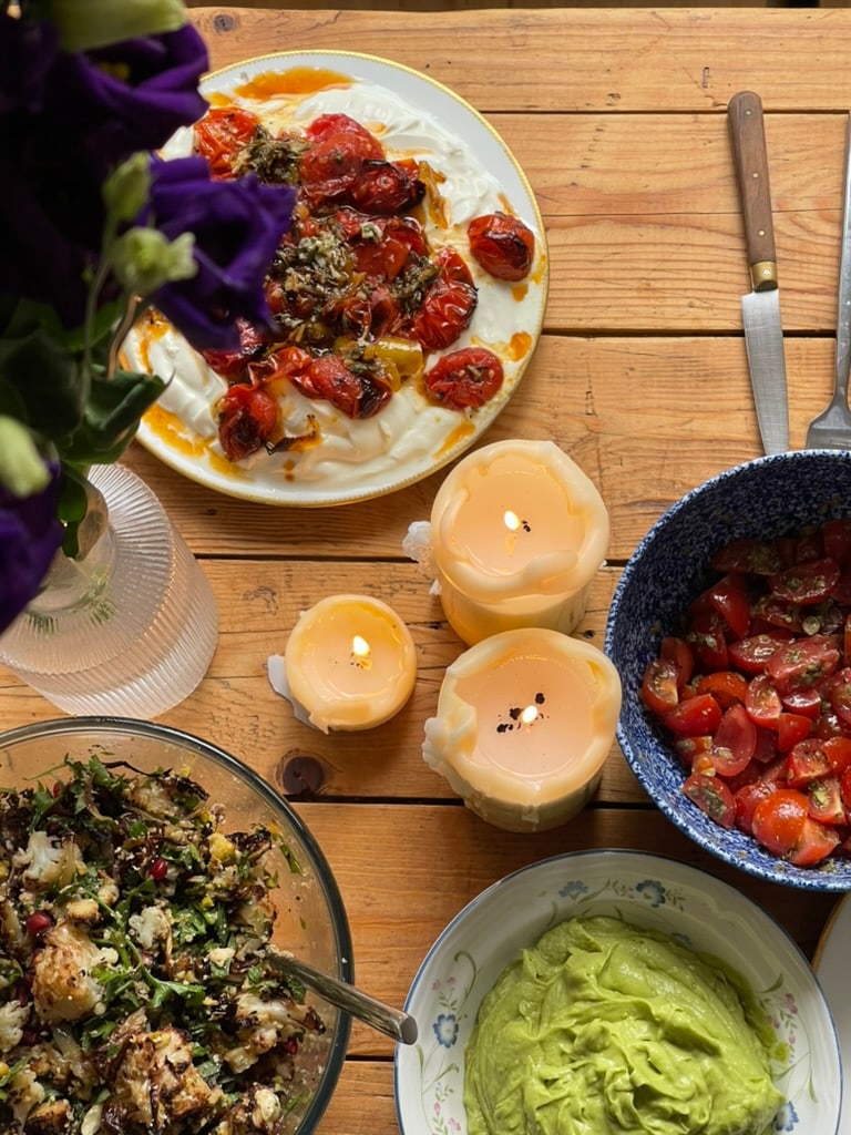 A long wooden table laid for friends - blistered tomatoes on labneh, charred grain salad, tomato salsa, avocado dip, candles burning low, deep purple lisianthus.